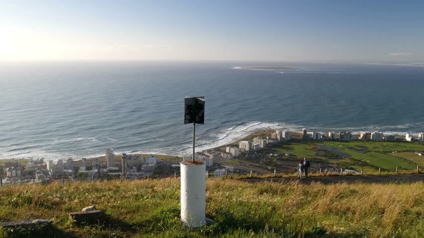 POV moving towards Signal Hill trig point marker & Green point aerial with coastline, ocean horizon, Robben Island, road with two people looking & pointing at view