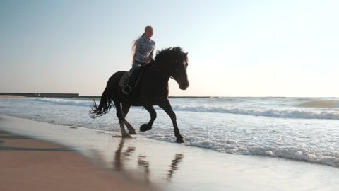 Beautiful young women riding dark horses at sea beach. Enjoying beautiful landscape.Galloping run in sunset or sunrise light and splashing water drops around.Riders running at a gallop.