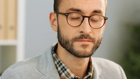 Close up face of an attractive caucasian man in spectacles, wearing a casual shirt and a grey shirt sitting at the office desk, looking and smiling at the camera.Indoor shot.