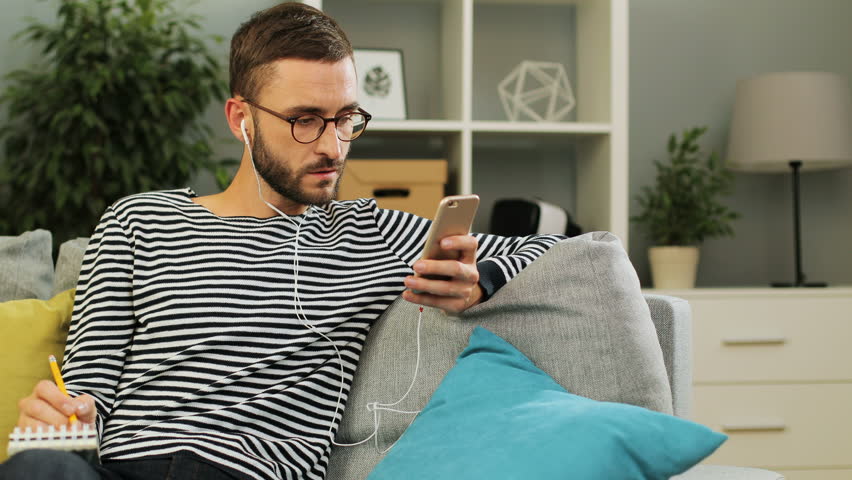Busy attractive man with earphones and cell phone in the hands taking notes to the copybook sitting on the couch with cushions on it at living room background. Indoor shot.