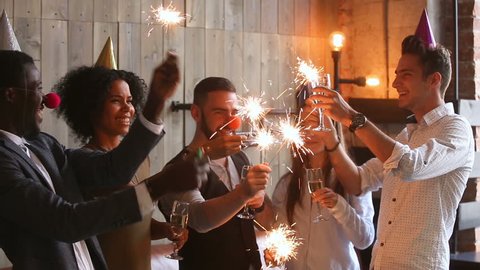 Multiracial young people wearing funny hats holding sparklers clinking glasses with champagne, happy african american and caucasian friends celebrating xmas, new year or birthday party indoors, cheers