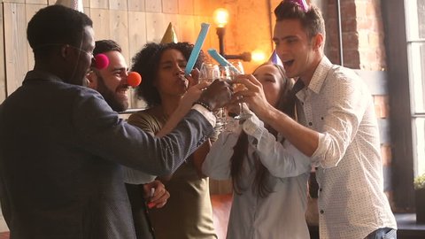 Multicultural young people in party hats clinking glasses celebrating holiday, diverse friends hanging out having fun holding champagne blowing party whistles at new years eve celebration indoors
