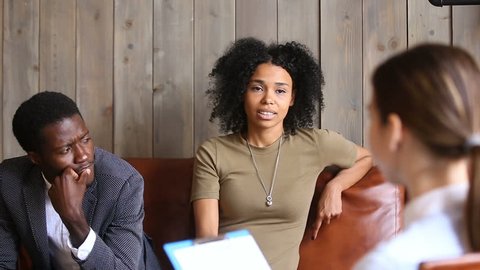 African couple sitting on couch at psychologists, black unhappy woman sharing marital problems talking to white counselor holding clipboard, family marriage counseling therapy session concept
