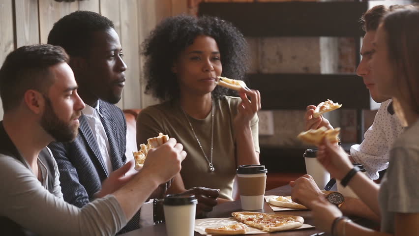 Multiracial happy young people eating pizza in pizzeria, black and white cheerful students enjoying meal dining sitting together at restaurant table, diverse hungry friends sharing lunch at meeting