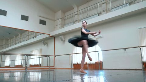 Ballerina in black tutu and pointe stretches on barre in ballet gym. Woman standing near bar and mirror, preparing for perfomance.