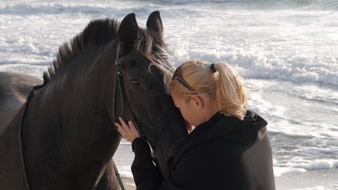 Young Blonde woman stroking and hugging horse. Slow motion. Beautiful lady with her black stallion on seashore enjoying nature. Love and friendship concept. Waves ocean background