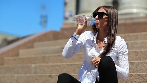 Young woman sits on the stairs and drinks mineral water from bottle. Beautiful girl with long hair in sunglasses drinks clean water. Girl opens plastic bottle of water and drinks soda water