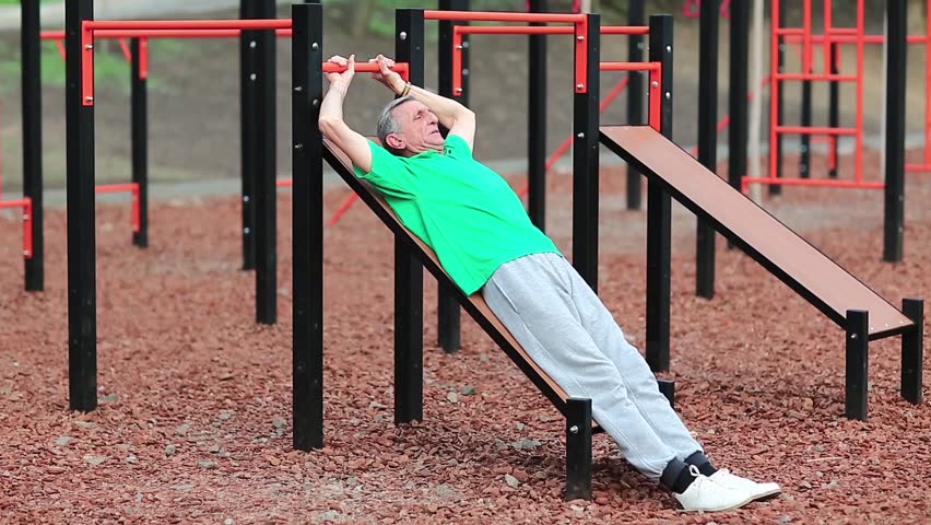 Senior man pumps abdominal muscles and burns up calories. Active elderly grey-haired athlete in green t-shirt doing physical exercise on sports ground, training in outdoor gym