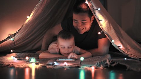 Father and son play and watching smartphone in children's room in tent with Christmas decorations