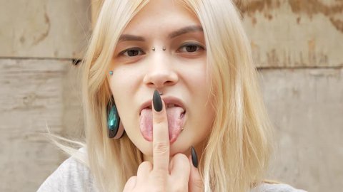 Close-up portrait of a young blonde with a split tongue, she shows a gesture and licks the middle finger. 4k. space for copying