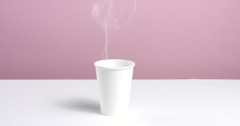 Hot water or coffee poured into a plain unlabeled paper cup on white table against pink wall, steam rising