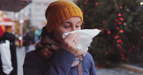 Sick Woman Blowing Her Nose Into Tissue Outdoors. SLOW MOTION 4K DCi. Young Female having cold or flu symptoms, standing in a busy city street. Autumn or winter cold weather.