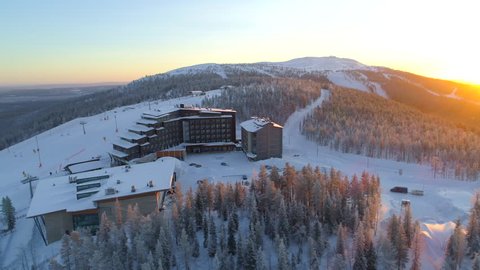 AERIAL: Flying around hilltop skiing resort surrounded by ski slopes in winter. Snowy hotel on top of ski tracks at golden winter sunset. Winter sun rising above quiet ski resort Levi in Finland