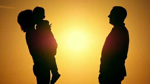 Silhouettes of happy family in slow motion embrace on sunset background. Mom holds the baby in her arms, Dad hugs and kisses them.