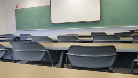 An empty lab in a computer science classroom. Chairs and tables sit in front of a chalkboard and projector screen. University hall represents lowered attendance rates, budget cuts and student debt.