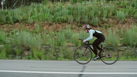 Tracking of tired female cyclist in protective helmet and professional sportswear riding bicycle along road surrounded by greenery