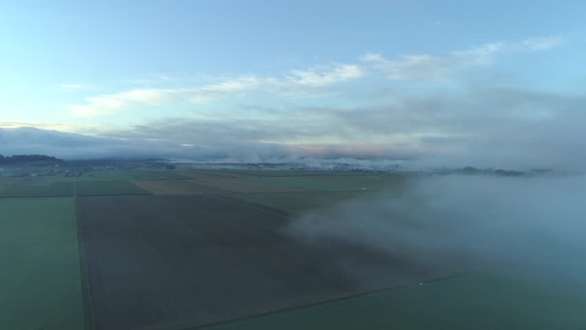 AERIAL: Thick fog rolling over vast rural meadow fields on cold winter morning. Cars driving through dense foggy clouds along the highway in rural countryside. Small industrial town dotted with houses
