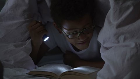 Child reading books at night under blanket, lighting himself with flashlight