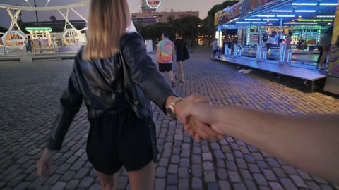 Attractive pretty couple spends date night at amusement park at night, girl or young teenager holds partner or boyfriend by hand and runs towards ferris wheel attraction, have fun and enjoy love mood