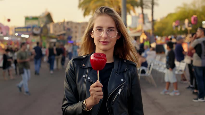 Beautiful blonde model, young woman in trendy cool black leather jacket walks around fair or carnival, inbetween attractions and rides and enjoys eats sugar candy apple in hard toffee caramel