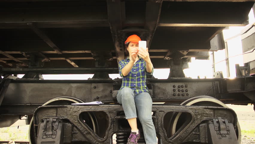 Young brunette woman railway worker in an orange helmet sits on the chassis frame of a freight wagon and making a video call using a camera on a white smartphone.