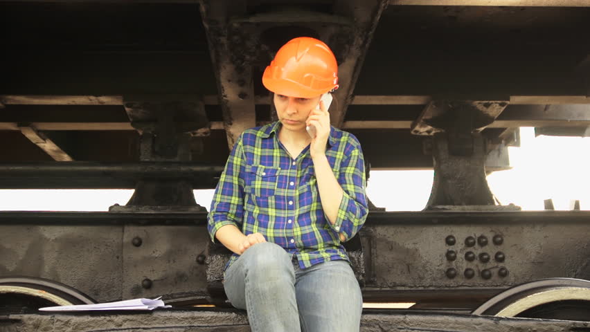 A young brunette woman railway worker in an orange helmet sits on the frame of the chassis of a freight car and phones a white smartphone.