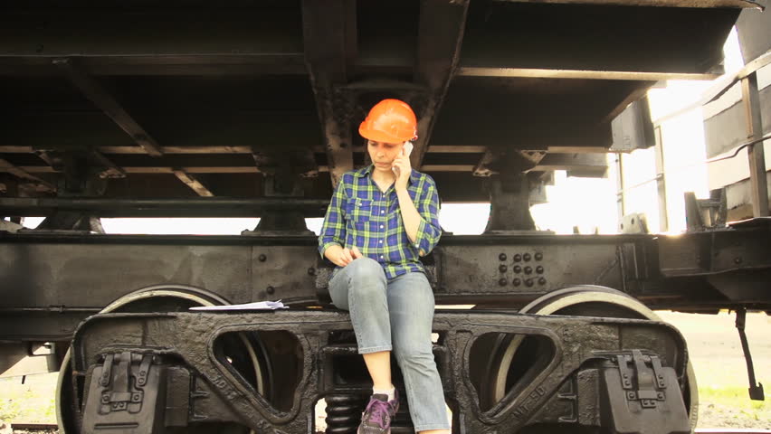 An alarmed woman, a railway worker, sits on the frame of the chassis of the freight wagon, shakes her foot, wipes sweat from her forehead under the orange helmet, speaks on the phone.