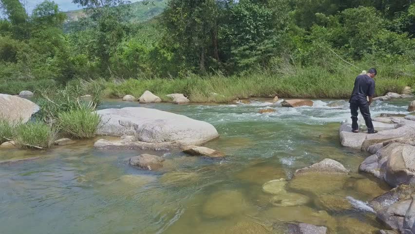 backside view local man stands on flat stone among river rapids and checks caught fish in net against green bank