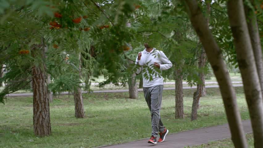 SLOWMO side view of African man in sportswear jogging in city park under green tree leaves