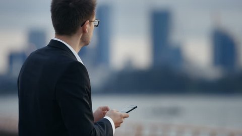 Young Businessman Uses Smartphone while Standing on the Street with Big City Skyscrapers View. Shot on RED EPIC-W 8K Helium Cinema Camera.
