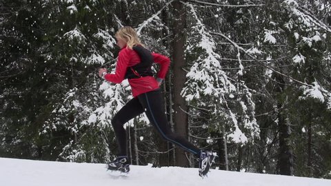 A women running uphill through a snowy forest in slow motion with crampons on her feet for traction.