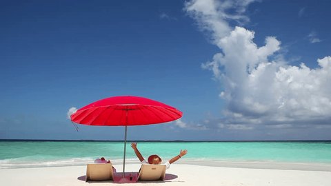 Rear view of happy couple with arms outstretched lying on back in deckchairs under the red parasol.White sand beach and turquoise water in Maldives beach