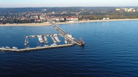 Sopot resort in Poland. Wooden pier (molo) with promenade, marina, yachts, pirate tourist ship, beach, old lighthouse, vacation infrastructure and far view of Gdynia. Aerial 4K video at sunrise.