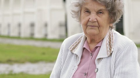 Black wristband. Mature elderly woman with gray hair aged 80s dressed in white jacket looks at the results of physical activity using a wristband fitness tracker outdoors in summer
