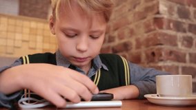 Closeup portrait of cute little kid uses smartphone with charge battery while sitting at table in restaurant or cafe. Boy drinks tea.  Real time full hd video footage.