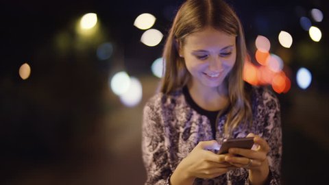 Attractive and young woman using a cell phone. She is checking mails, chats or the news online. Night. Traffic passing by.
