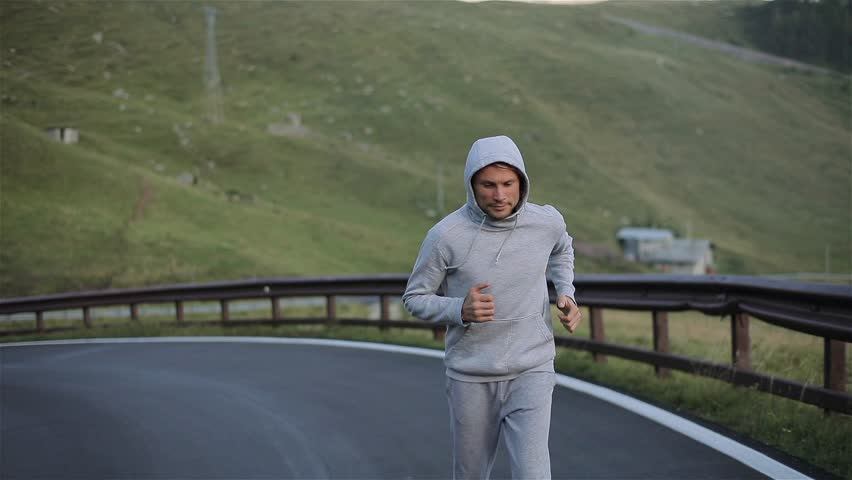 Man jogging on mountain road close up slow motion looking at camera front side view. Morning jogger moves hands running up valley hill in grey hood sportswear. Ambition goals training success concept Royalty-Free Stock Footage #31615897