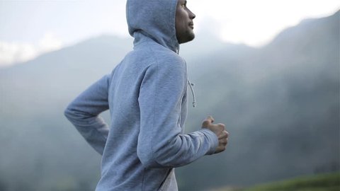 Man jogging on mountain road close up slow motion looking at camera front side view. Morning jogger moves hands running up valley hill in grey hood sportswear. Ambition goals training success concept