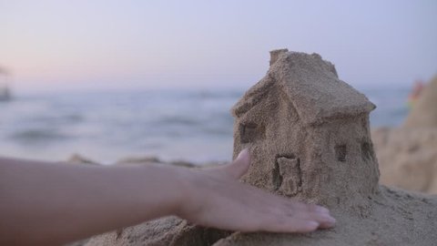 Small house with a roof made of sand on the beach at sunset. Property insurance, house protection or saving and security concept. Home under woman's care. Protecting gesture of young girl.