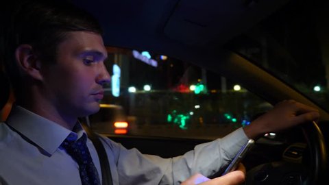 Texting with a smart phone in car. a young businessman is driving a car around the night city and uses his smartphone. 4k, slow motion