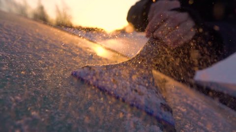 SLOW MOTION CLOSE UP DOF: Person cleaning morning frost off a car window at golden sunrise. Hand scraping hoarfrost off a frozen car windshield, snowflakes flowing over the sun. Scraper removing frost