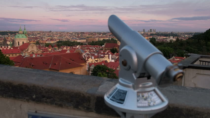 Camera slides past tourist telescope to show rooftops and skyline of Prague at sunset