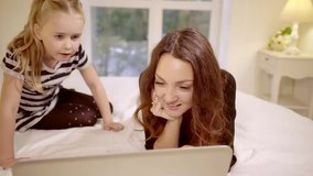 mother and her cute little daughter are watching funny video on a laptop computer, lying on a bed