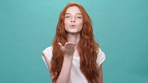 Smiling ginger woman in t-shirt sends air kiss at the camera over turquoise background