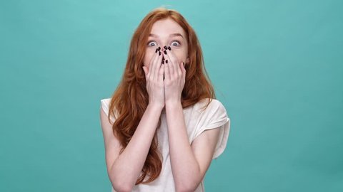 Shocked happy ginger woman in t-shirt covering mouth and looking at the camera over turquoise background
