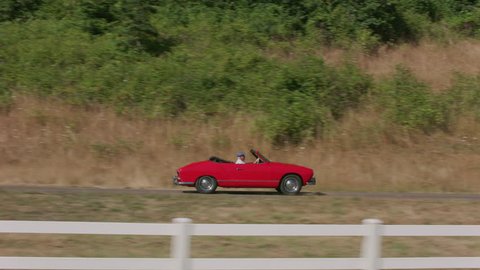 Tracking shot of man driving classic convertible car down fence lined driveway. Fully released for commercial use.