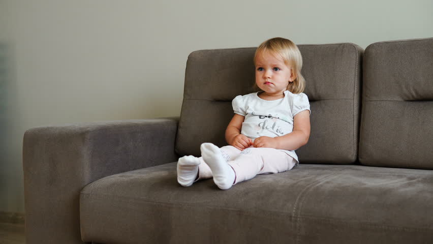 A child with a serious face sits on the couch. Little girl is watching TV alone. The child bites his lip. A cute little girl is watching a scary movie. The child is scared of what he saw on TV. | Shutterstock HD Video #31658773