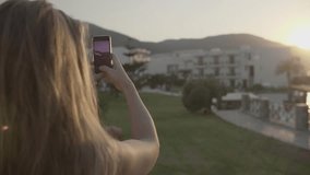 Young blonde woman is taking photo with smartphone and looking at panorama view of resort at sunset. Attractive girl takes pictures on cell phone. Vacation and destination concept.