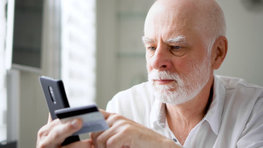 Handsome bearded smiling senior man sitting at home. Shopping online with credit card on cellphone. Concept of technology use by older people. Active modern life after retirement | Shutterstock HD Video #31667281