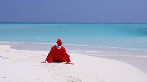 Man in Santa Claus Hat sitting and wag hands on sandy beach. Christmas vacation on Maldivian islands